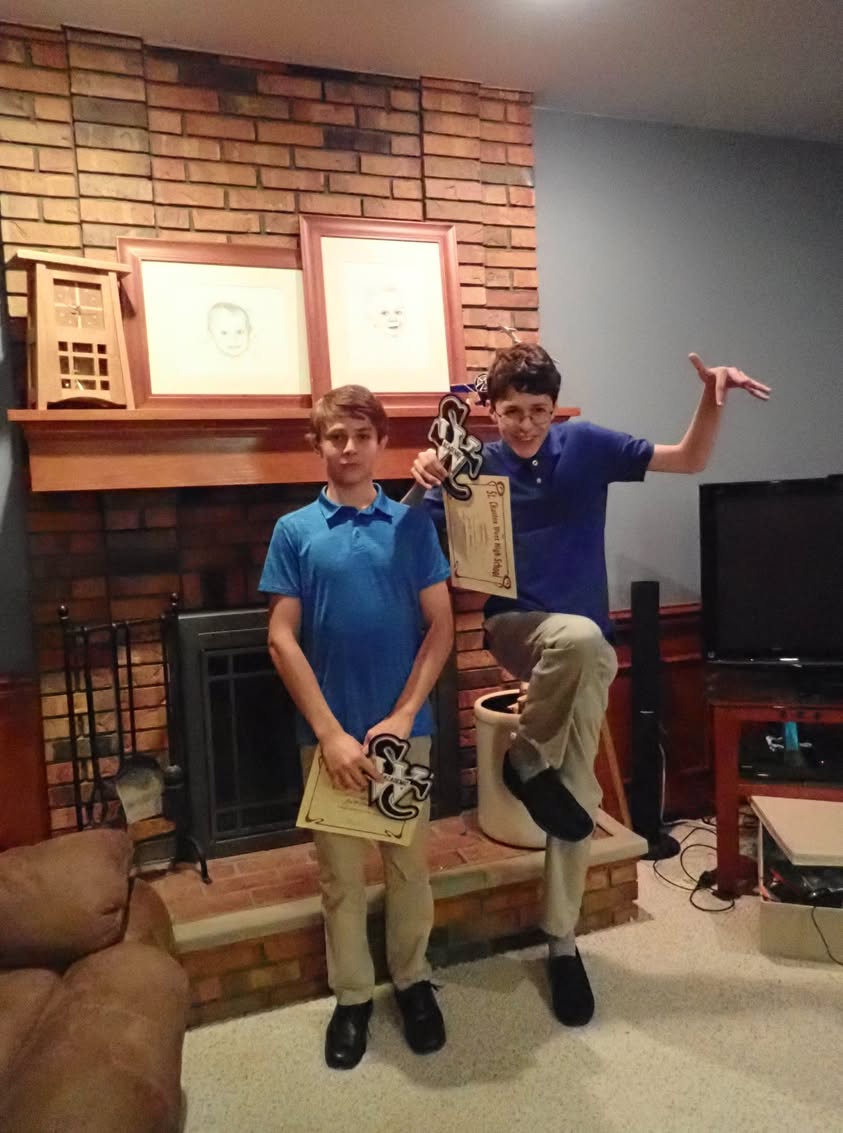 Two boys hold academic letter awards in front of a brick fireplace, with one posing playfully.