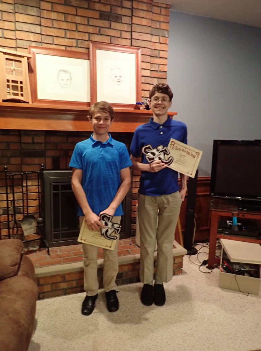 Two boys stand together holding academic letter awards in front of a brick fireplace.