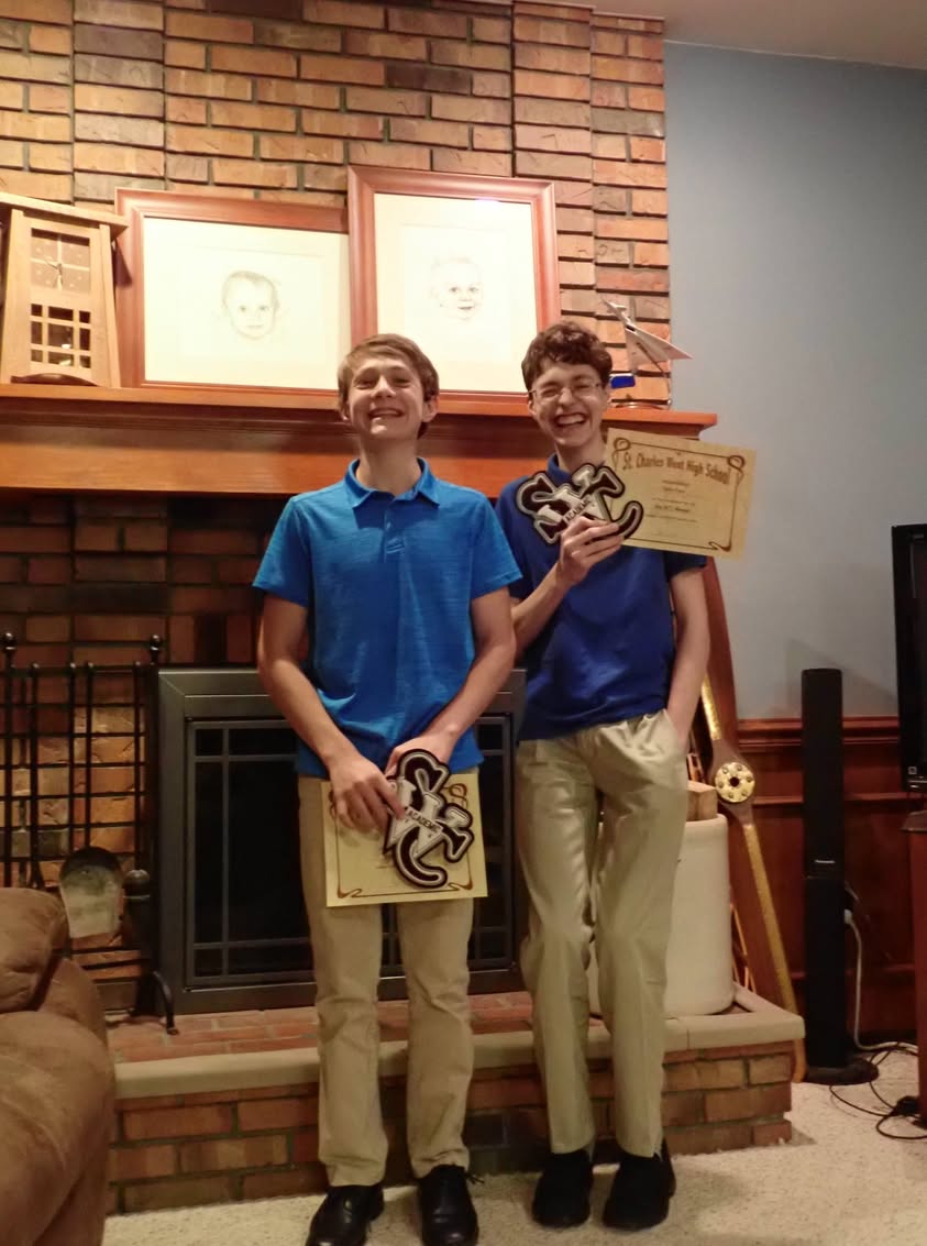 Two boys smile and laugh while holding academic letter awards in front of a brick fireplace.
