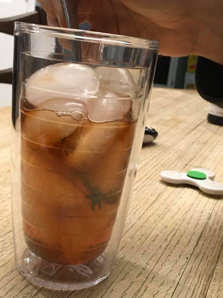 A clear cup of iced tea with a small clay figure submerged inside.