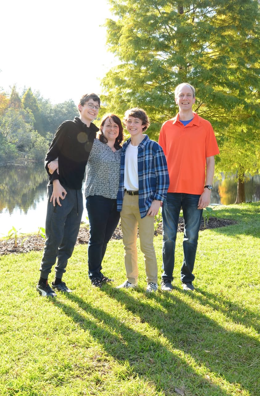 Family of four standing together outdoors beside a pond on a sunny day.