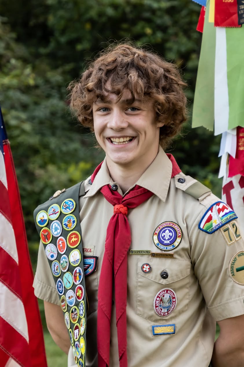 A smiling Eagle Scout in uniform standing outdoors beside an American flag.