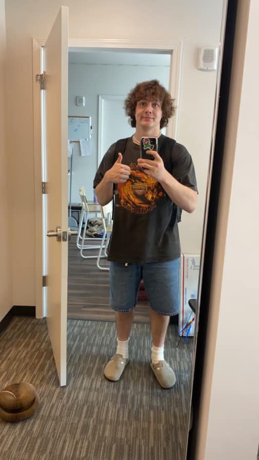 A young man taking a mirror selfie in a room, giving a thumbs-up while wearing a dark T-shirt and denim shorts.