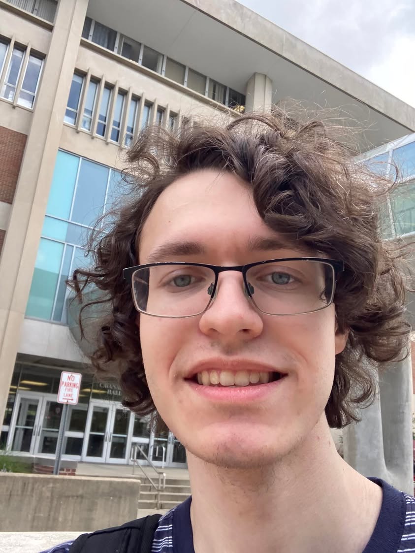 Young man with glasses taking an outdoor selfie in front of a campus building.