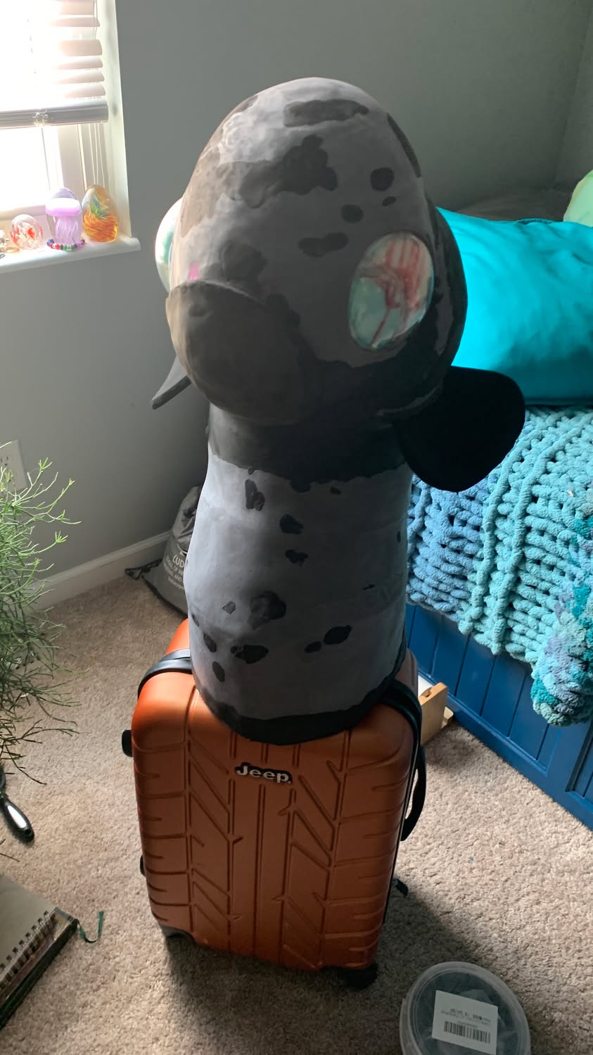 Large gray handmade creature head with round side pieces resting on top of an orange case.