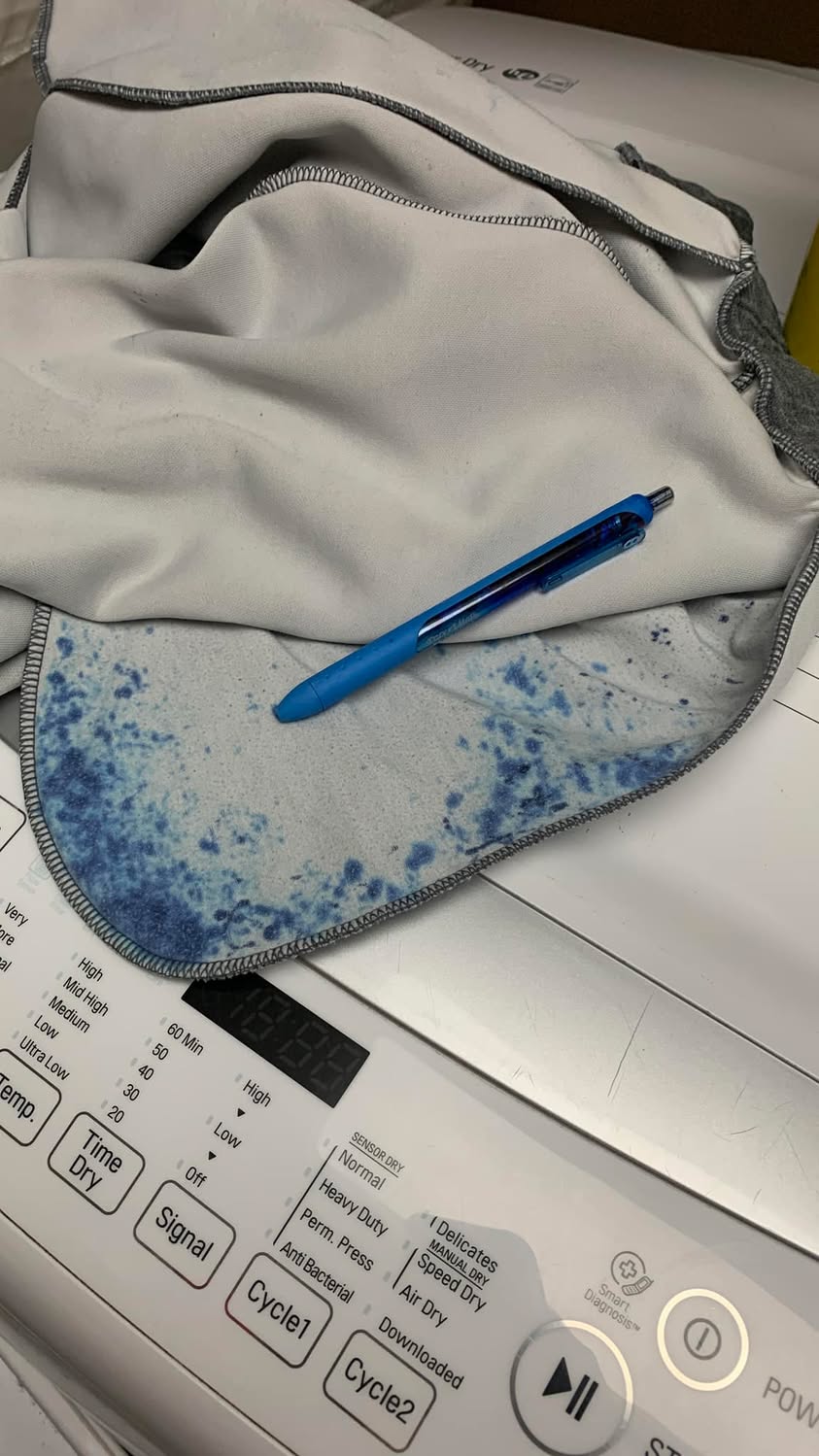 Laundry stained with blue ink on top of a dryer, with a blue pen resting on the fabric.