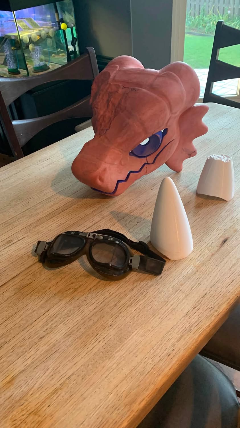 A large orange handmade creature head on a kitchen table with goggles and two white cone-shaped pieces.