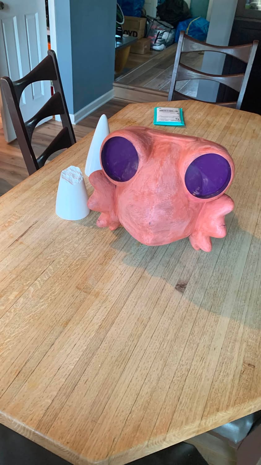 Front view of a large orange handmade creature head with purple eye openings and white cone-shaped pieces on a kitchen table.